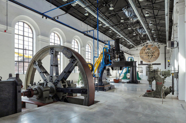 Wnętrze przemysłowego muzeum z dużym kołem maszynowym, architektonicznymi detalami w postaci białych arkadowych okien i współczesnymi instalacjami